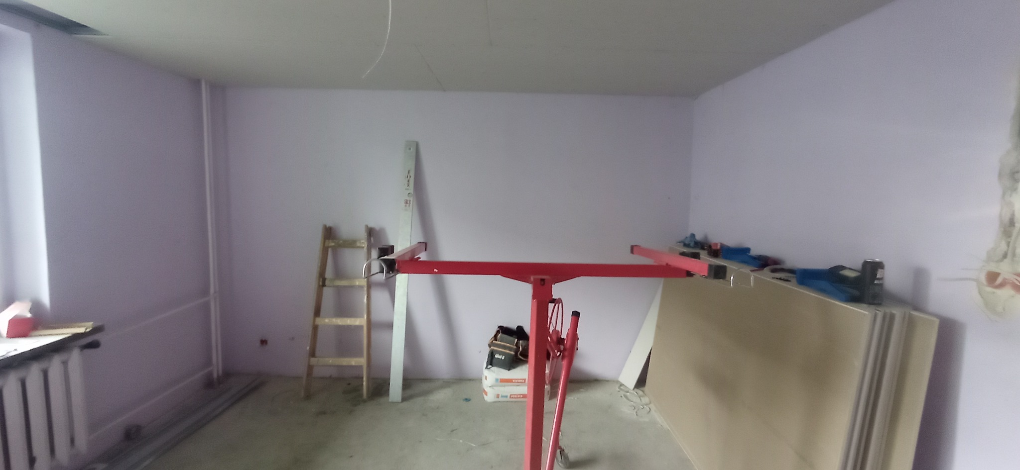 Wnętrze pokoju w trakcie prac wykończeniowych. Widoczny podnośnik do płyt, drabina, płyty i narzędzia. Ściany pomalowane na jasny kolor.