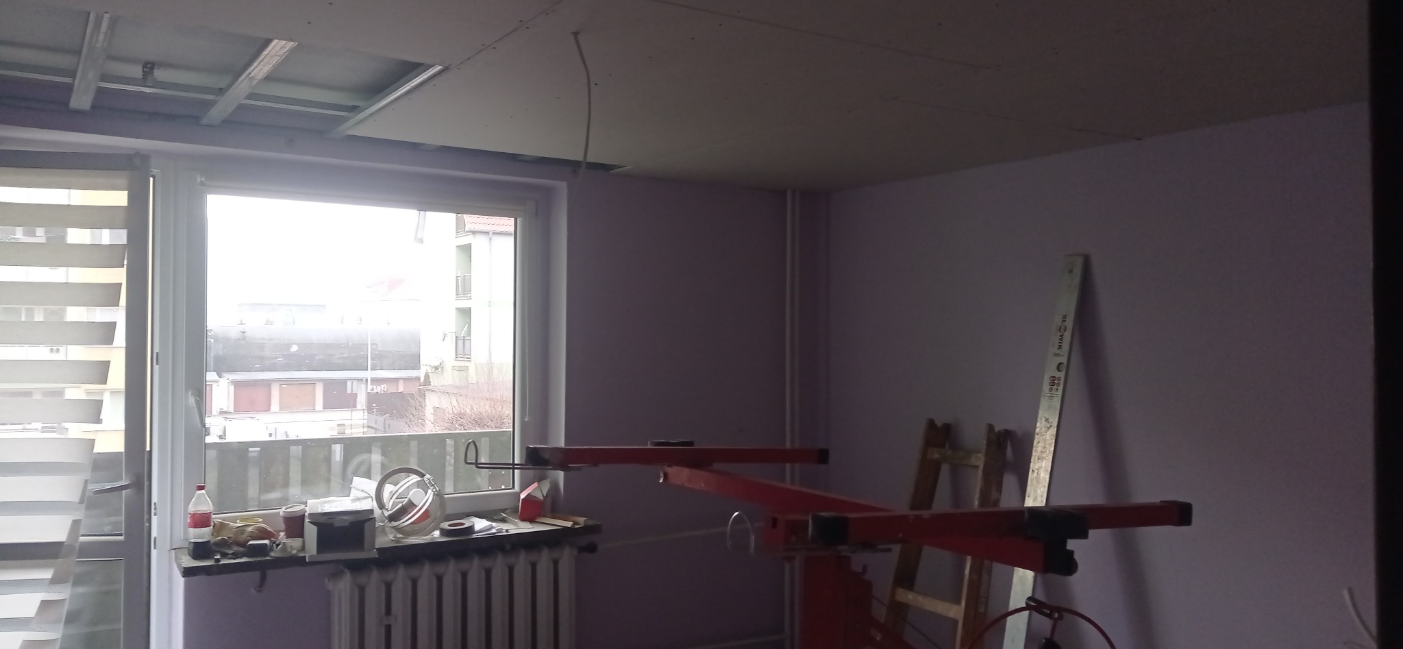 Montaż sufitu podwieszanego z płyty gipsowej w pokoju. Widoczny stelaż, częściowo zamontowana płyta, podnośnik i drabina. Okno z widokiem na budynki.