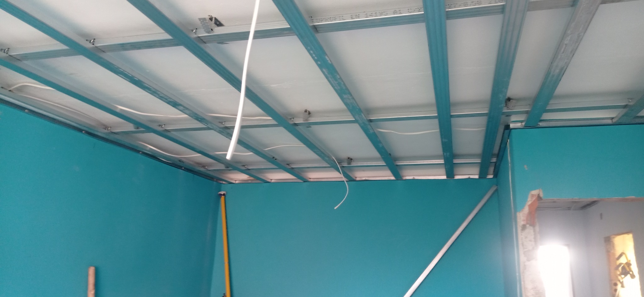 Montaż sufitu podwieszanego z płyt gipsowych na metalowej konstrukcji w pomieszczeniu z turkusowymi ścianami. Widoczne przewody elektryczne.