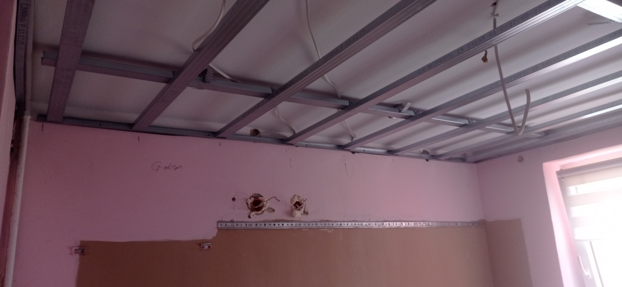 Montaż metalowej konstrukcji sufitu podwieszanego z płyt gipsowo-kartonowych w pomieszczeniu z różowymi ścianami i widocznymi instalacjami elektrycznymi.