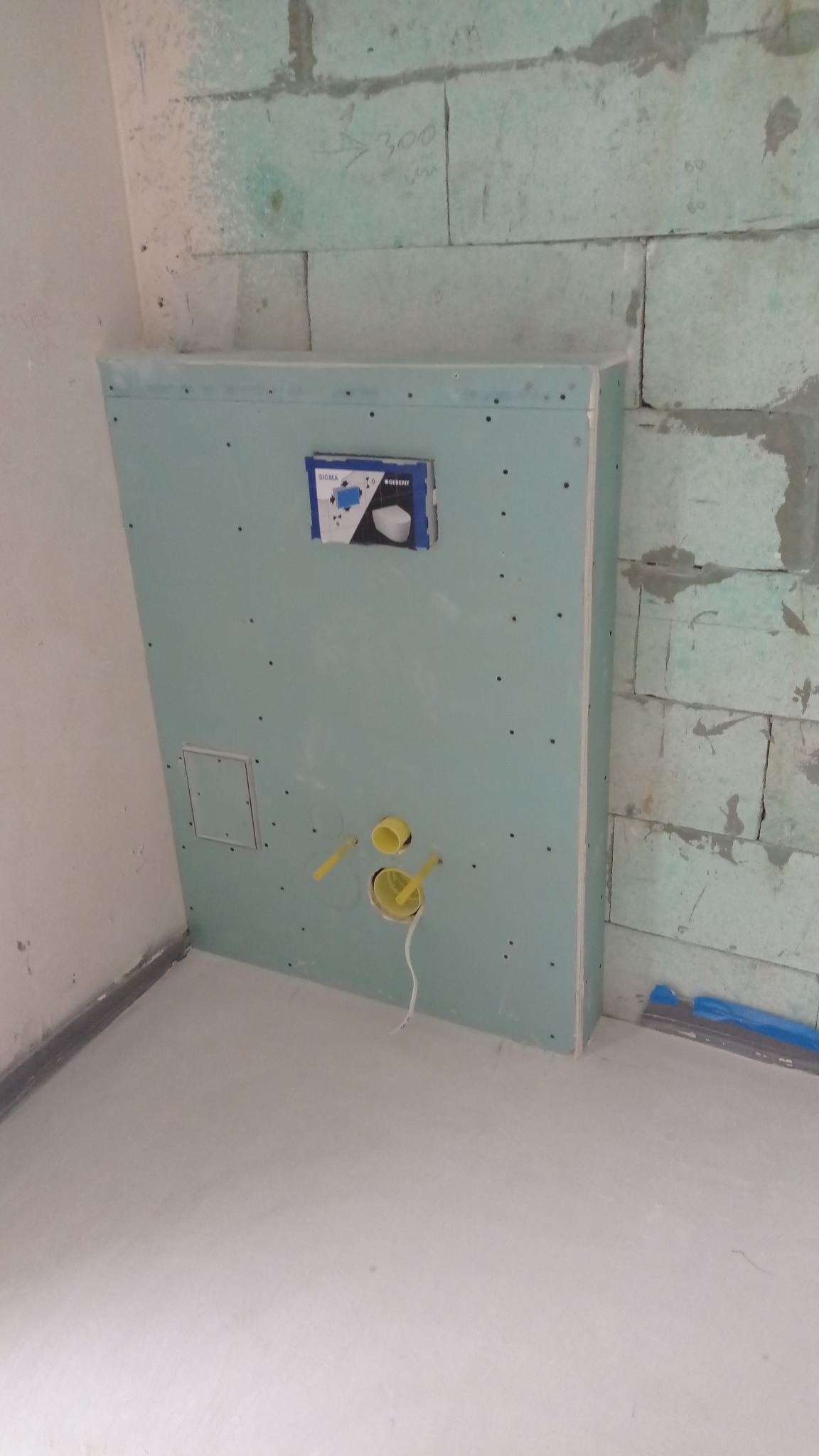 Ścianka z płyty GK z instalacją podtynkową WC w trakcie montażu, widoczne żółte rury i otwór rewizyjny. Ściana z betonu komórkowego w tle.