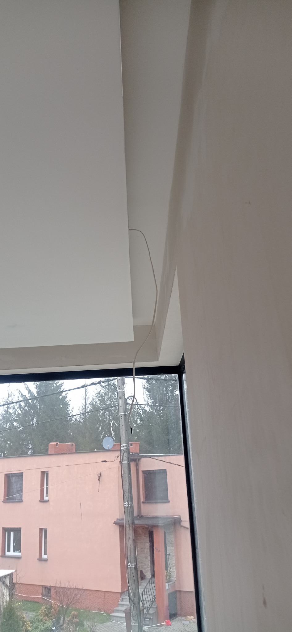 Wykończony fragment sufitu podwieszanego z widocznym kablem, ściana po szpachlowaniu, okno z widokiem na budynek i drzewa. Ujęcie z dołu, z bliska.