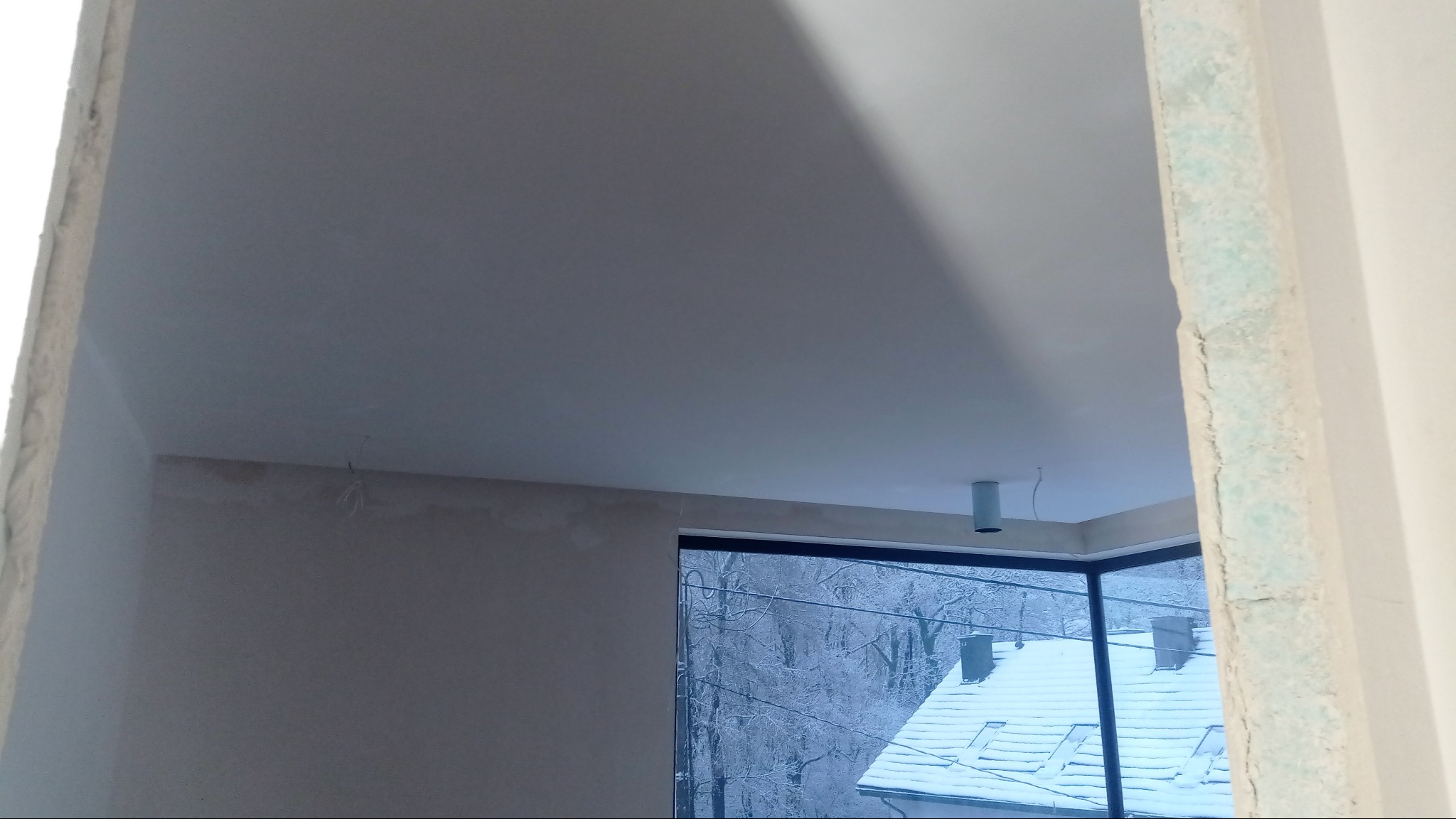 Wnętrze w trakcie prac wykończeniowych: gładka, jasna ściana i sufit, widok na zaśnieżony krajobraz przez okno. Fragment ościeżnicy z widocznymi śladami obróbki.