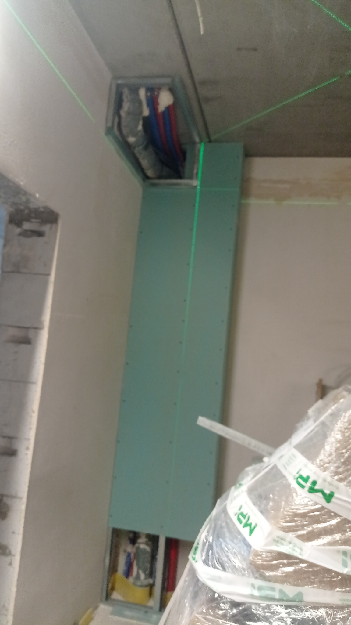 Montaż płyty G-K na ścianie z widocznymi przewodami w otworze, wyznaczony pion lasera. Prace wykończeniowe w surowym wnętrzu.