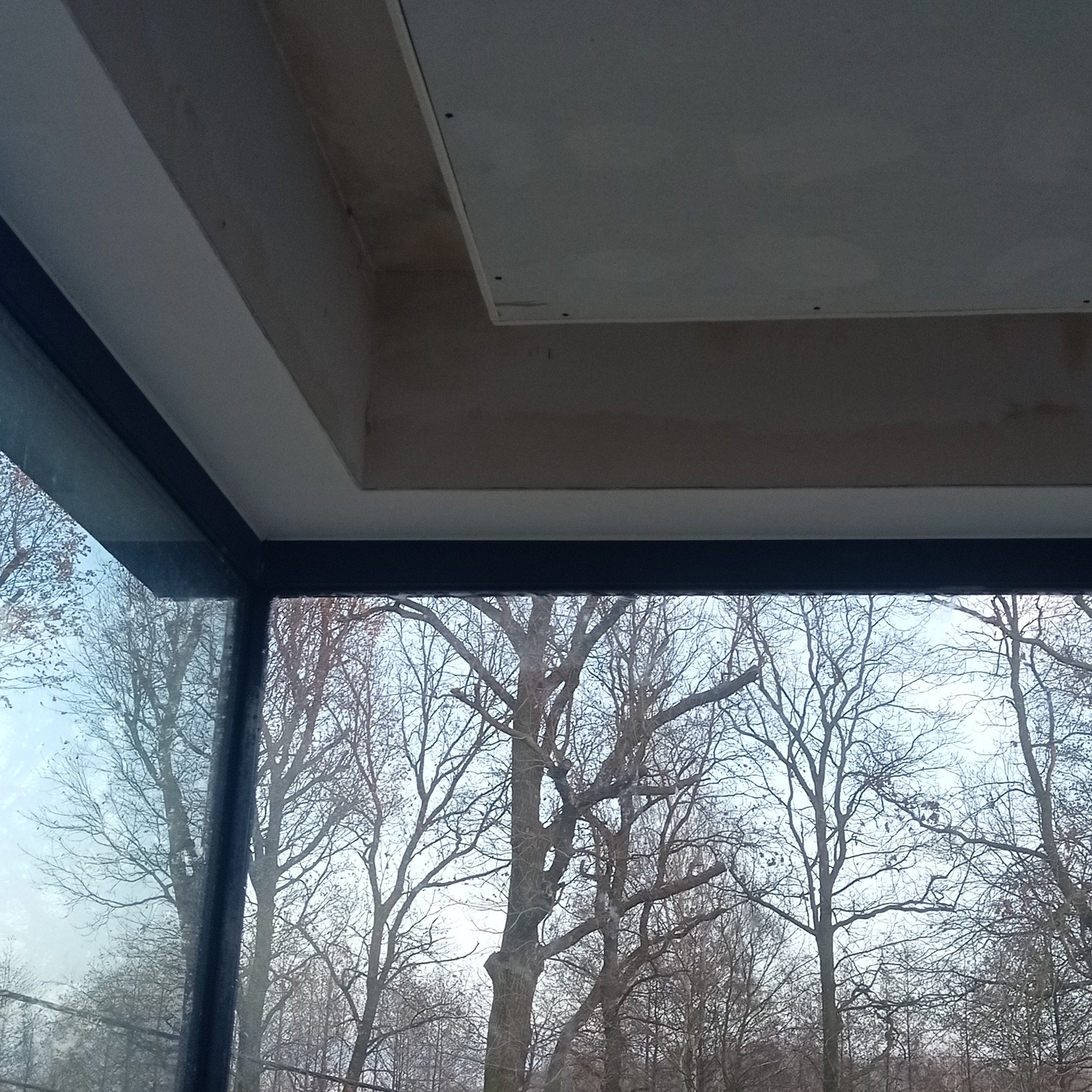 Narożnik okna z widokiem na drzewa, fragment sufitu z płytą kartonowo-gipsową w trakcie montażu. Widoczne łączenia płyt i niedoskonałości powierzchni.