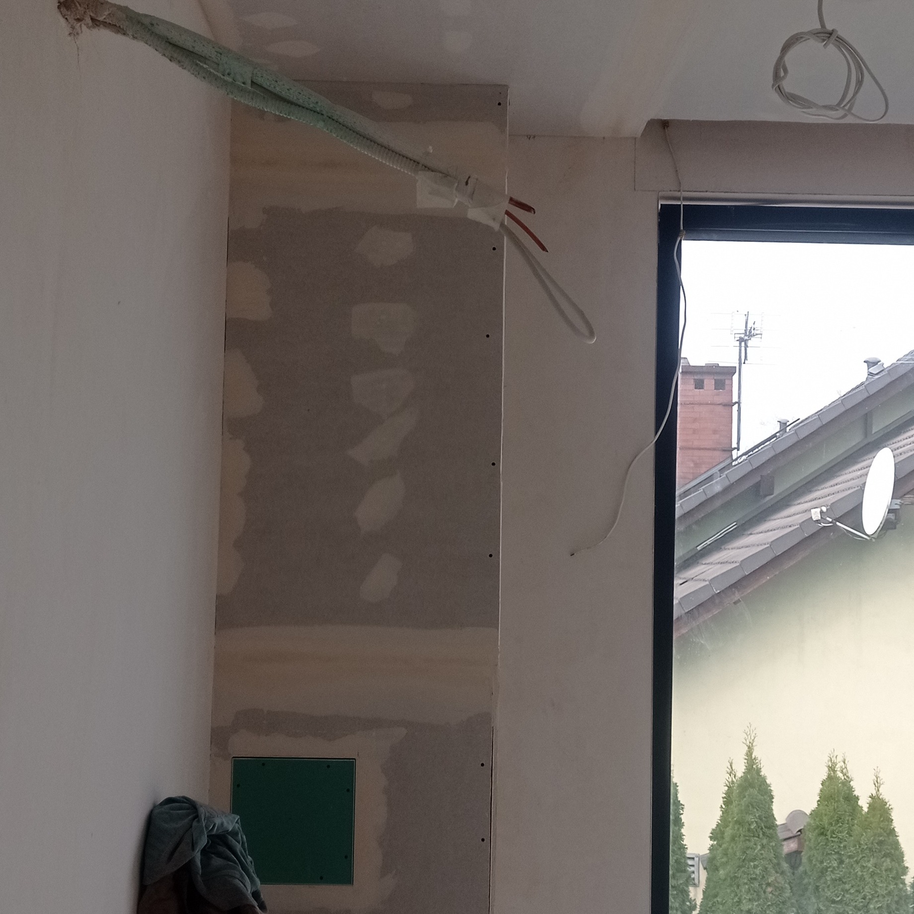 Ściana z płyt karton-gips, widoczne kable i instalacje, szpachlowane łączenia, okno z widokiem na domy. Prace wykończeniowe w trakcie realizacji.