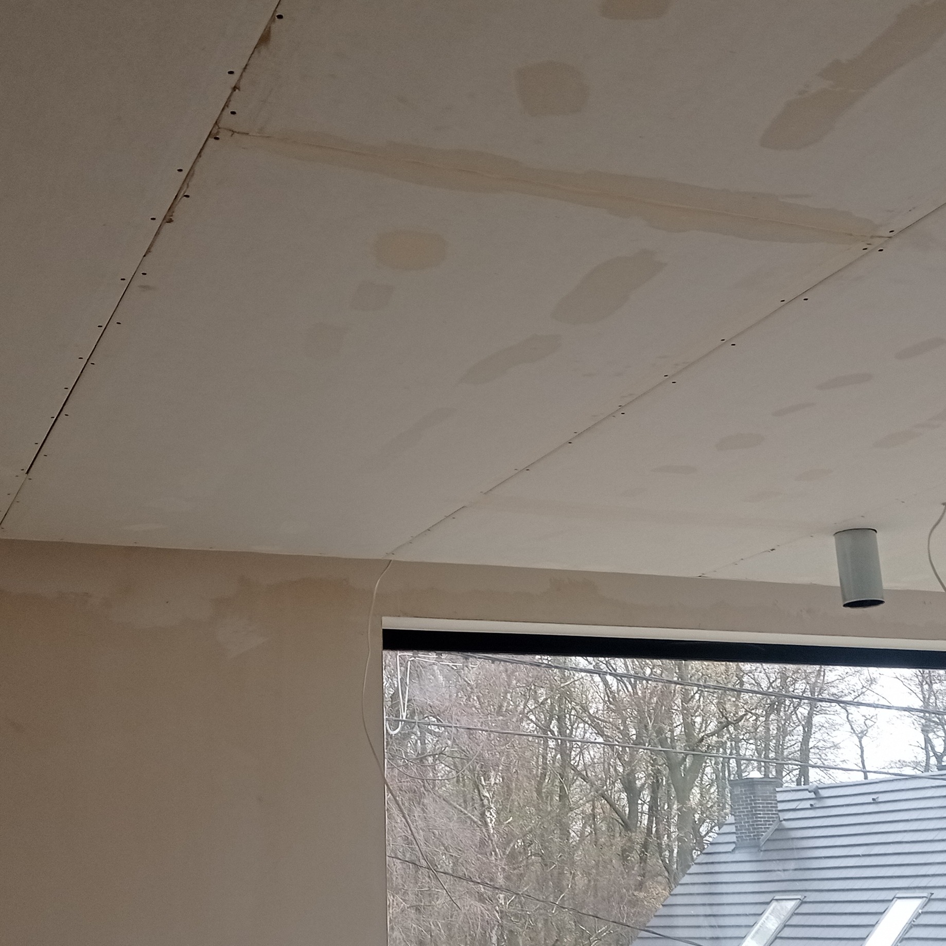 Sufit z płyt kartonowo-gipsowych, widoczne łączenia i ślady szpachlowania, fragment okna z widokiem na drzewa w tle. Oświetlenie sufitowe w prawym górnym rogu.