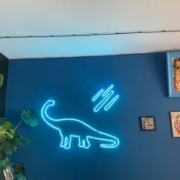 Niebieski neon w kształcie dinozaura Brontozaura na ciemnoniebieskiej ścianie, uzupełniony abstrakcyjnym neonowym wzorem i roślinami doniczkowymi w rogu pokoju.