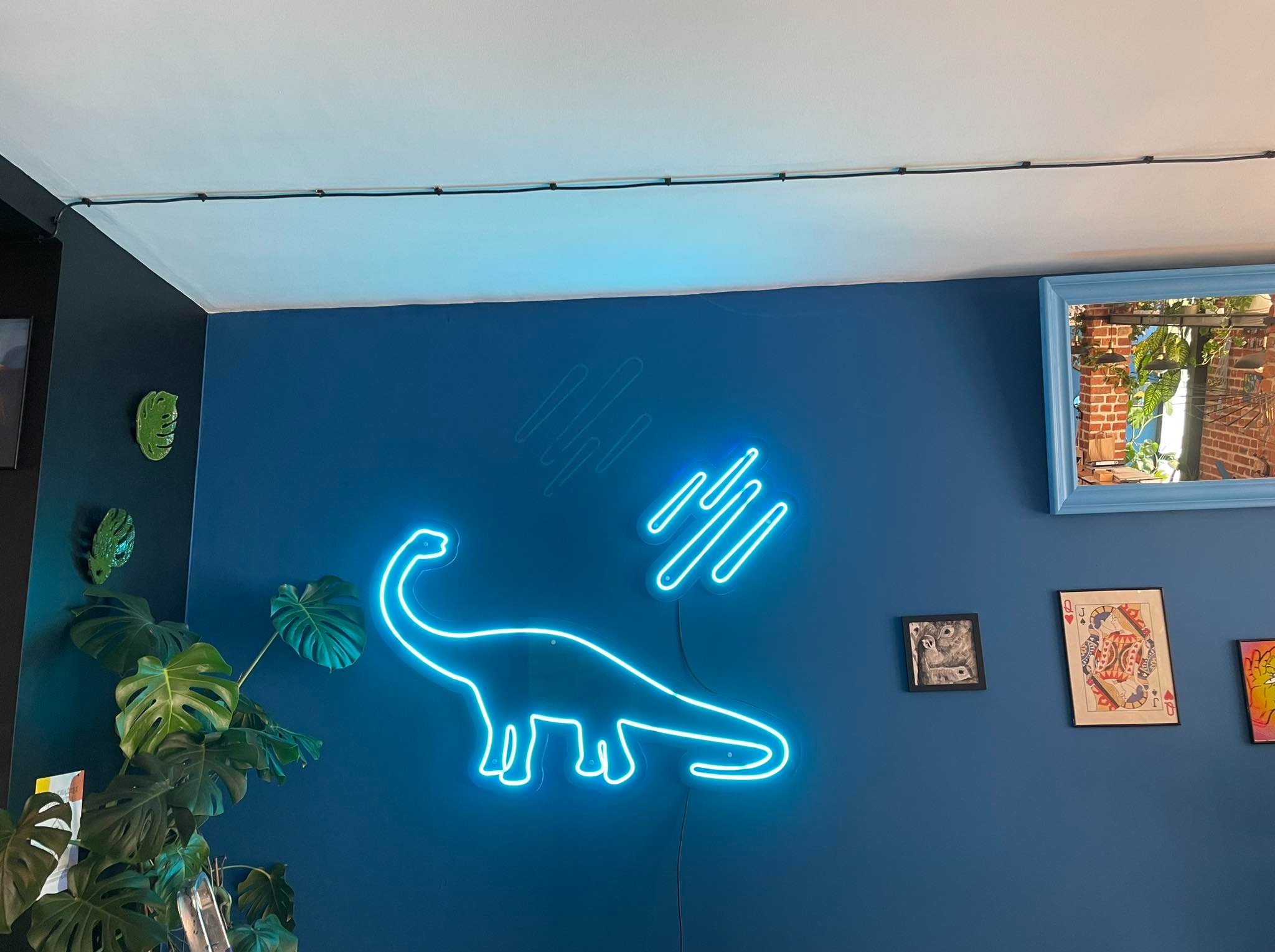 Niebieski neon w kształcie dinozaura Brontozaura na ciemnoniebieskiej ścianie, uzupełniony abstrakcyjnym neonowym wzorem i roślinami doniczkowymi w rogu pokoju.