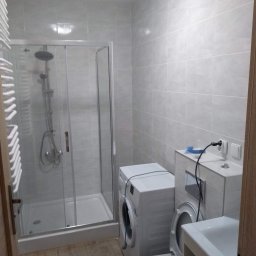 AleksandrijaBud Sp z OO - Jasna łazienka z kabiną prysznicową, pralką i umywalką z szafką, wykończona płytkami w odcieniach szarości, widok na fragment toalety z otwartą klapą sedesową.