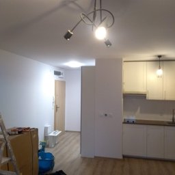 AleksandrijaBud Sp z OO - Wnętrze mieszkania w trakcie prac wykończeniowych: widoczna drabina, narzędzia, kartony, świeżo pomalowane ściany i zabudowa kuchenna.