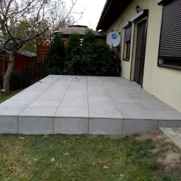 AleksandrijaBud Sp z OO - Wykonany z szarych płyt taras przylegający do budynku z beżową elewacją, widoczne rolety zewnętrzne i antena satelitarna, w tle drzewa i trawnik.