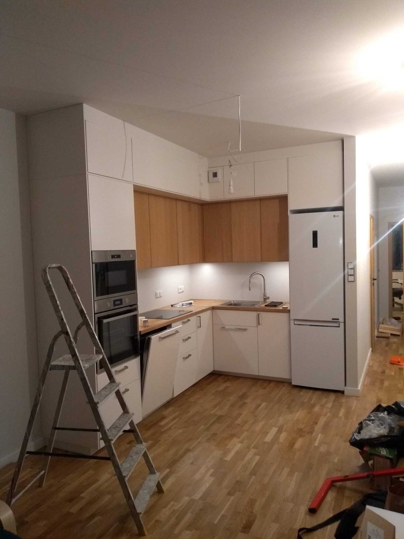 Ukończona kuchnia w stylu skandynawskim z białymi i drewnianymi szafkami, widok na zabudowane sprzęty AGD i drabina malarska w rogu.