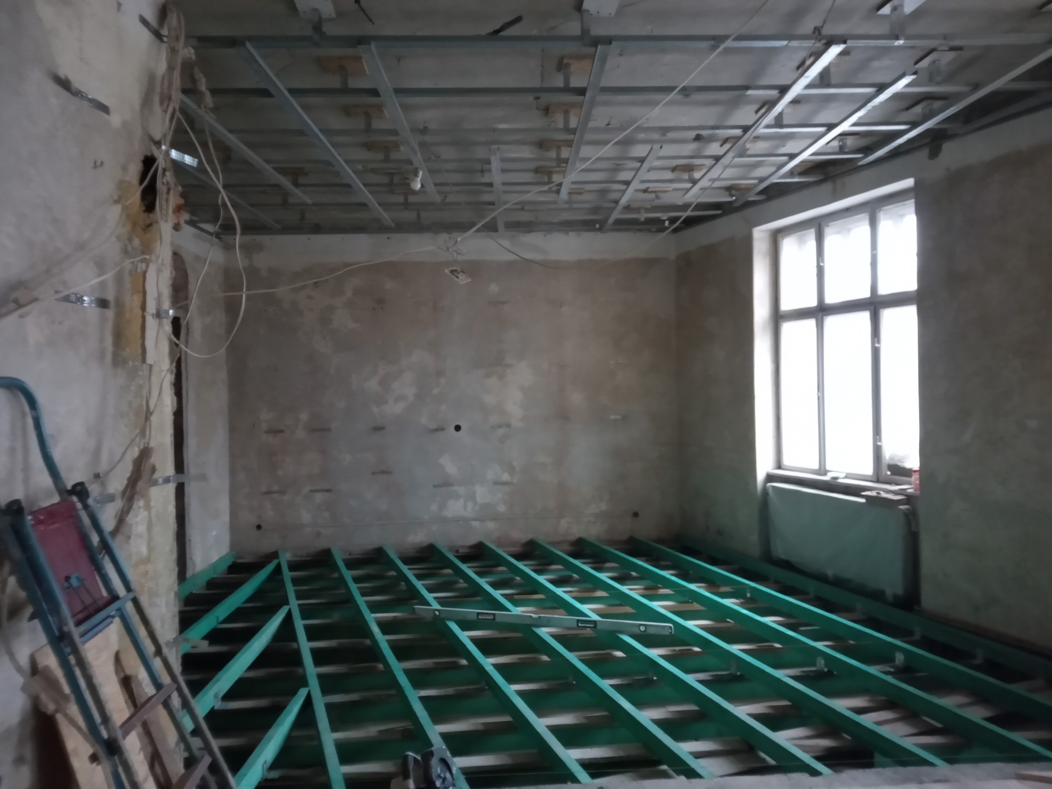 Wnętrze w trakcie remontu: zielona konstrukcja podłogowa, metalowy stelaż sufitowy, surowe ściany, drabina oparta o ścianę, poziomica na podłodze, duże okno z parapetem.
