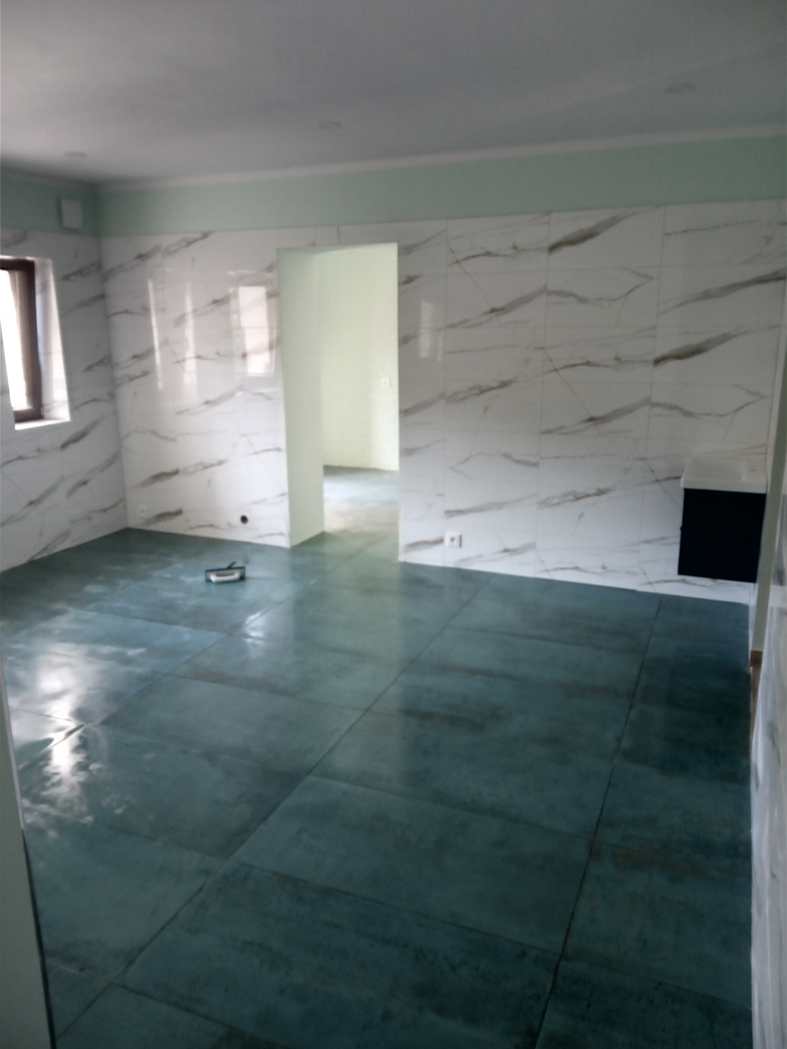 Świeżo wykafelkowane pomieszczenie z błyszczącymi, turkusowymi płytkami podłogowymi i białymi płytkami ściennymi z marmurkowym wzorem; widoczny otwór drzwiowy do kolejnego pomieszczenia.