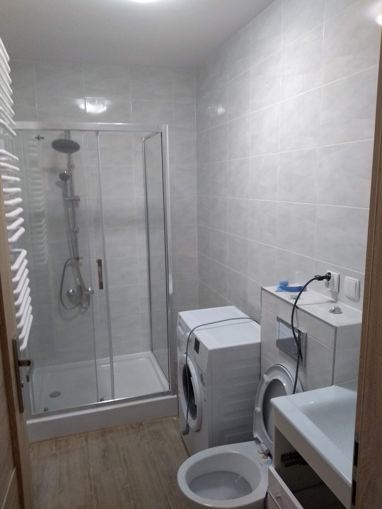 Jasna łazienka z kabiną prysznicową, pralką i umywalką z szafką, wykończona płytkami w odcieniach szarości, widok na fragment toalety z otwartą klapą sedesową.