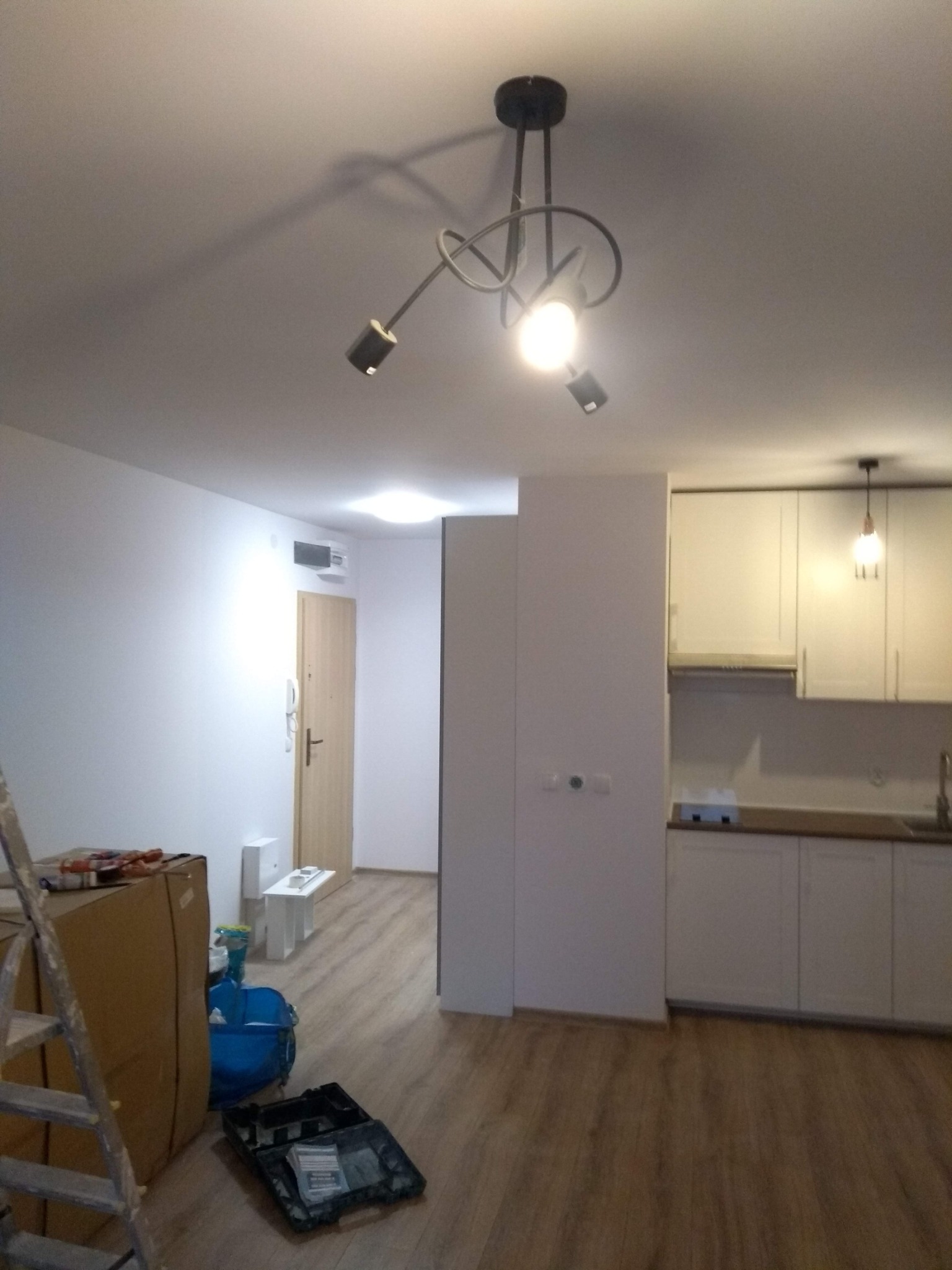 Wnętrze mieszkania w trakcie prac wykończeniowych: widoczna drabina, narzędzia, kartony, świeżo pomalowane ściany i zabudowa kuchenna.