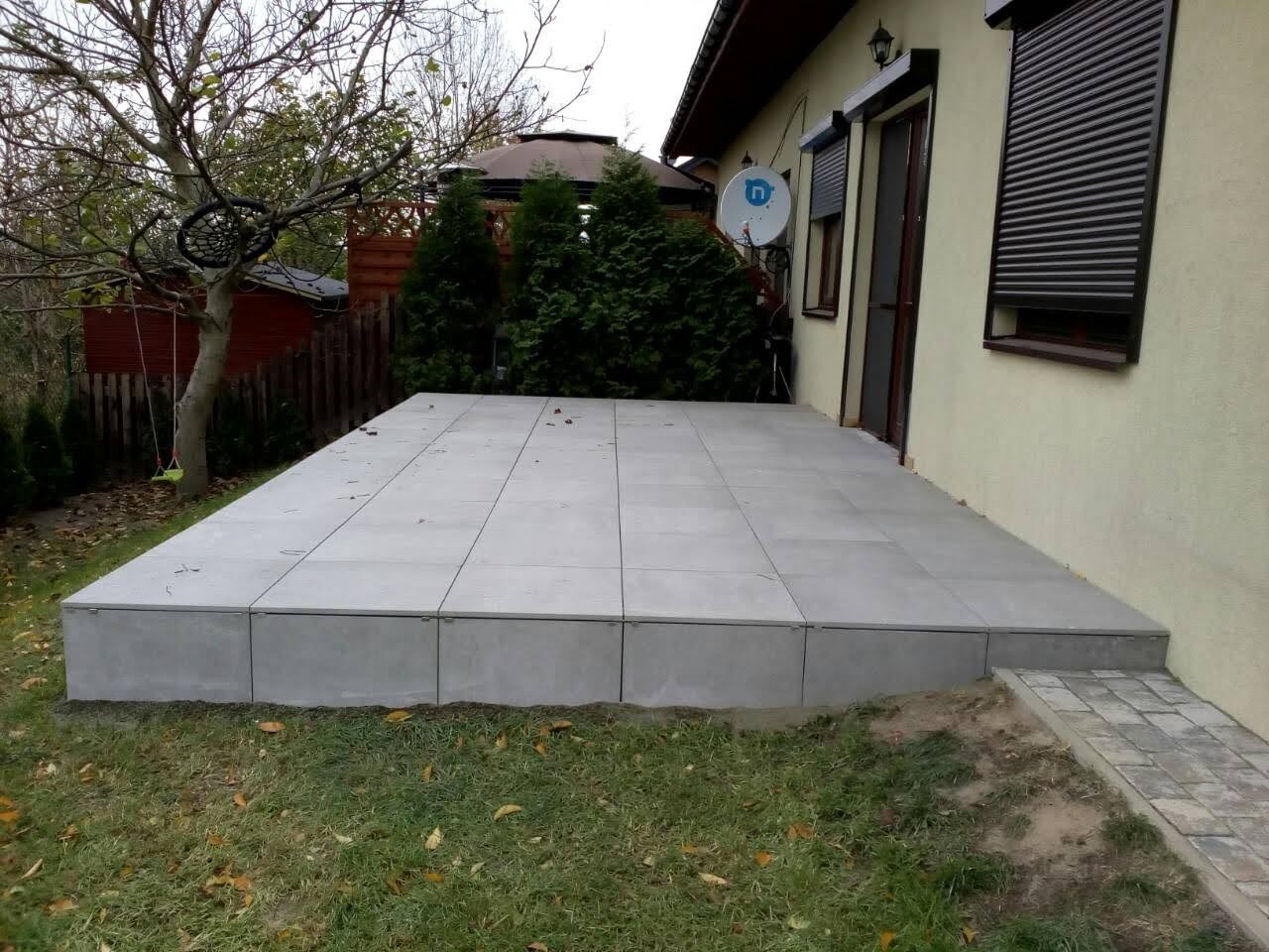 Wykonany z szarych płyt taras przylegający do budynku z beżową elewacją, widoczne rolety zewnętrzne i antena satelitarna, w tle drzewa i trawnik.