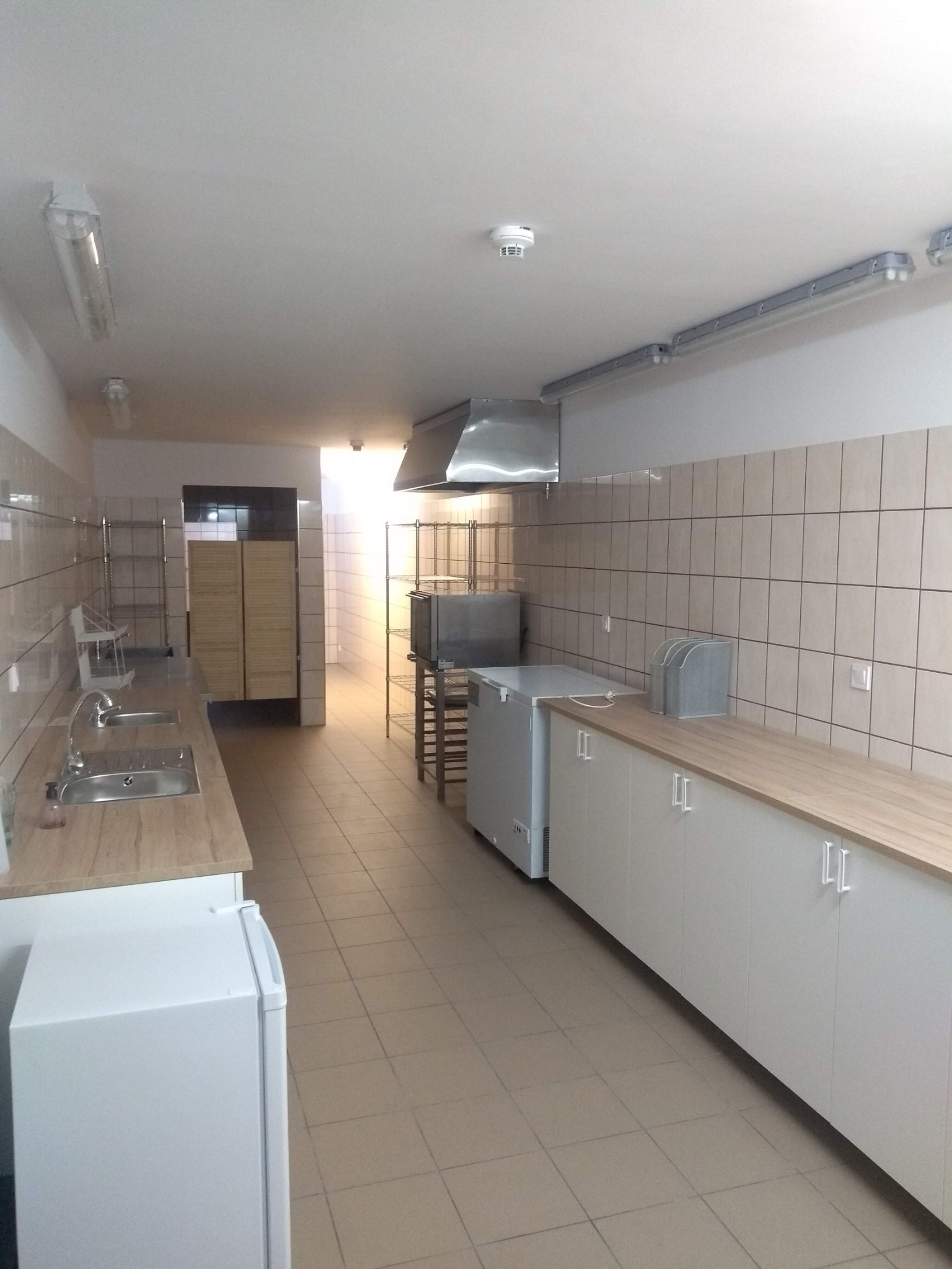 Wyposażone wnętrze kuchni przemysłowej z okapem, szafkami, blatem i lodówkami w jasnej tonacji.