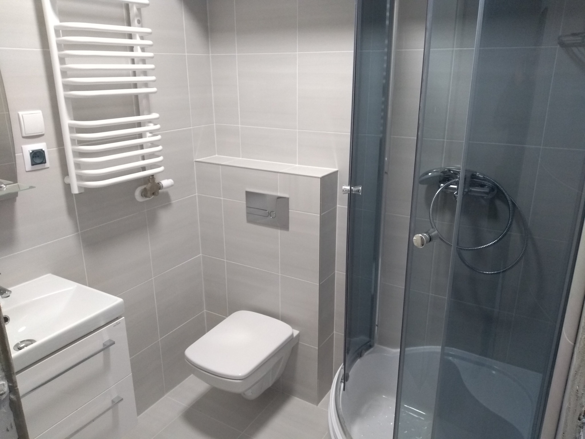 Nowoczesna łazienka z szarymi płytkami, wiszącą toaletą, kabiną prysznicową z ciemnymi szybami i białym grzejnikiem drabinkowym.