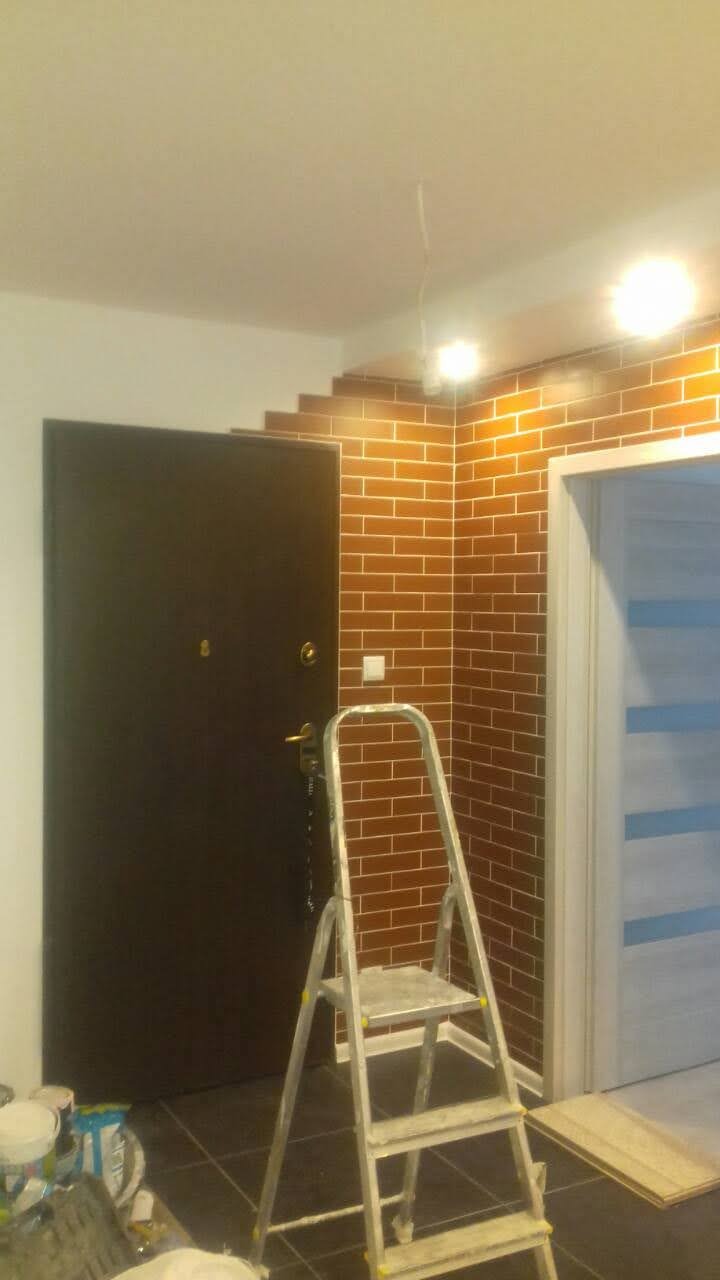 Korytarz w trakcie prac wykończeniowych: ściana wyłożona brązowymi płytkami imitującymi cegłę, metalowa drabina, czarne drzwi wejściowe, białe drzwi wewnętrzne z niebieskimi paskami, widoczne...