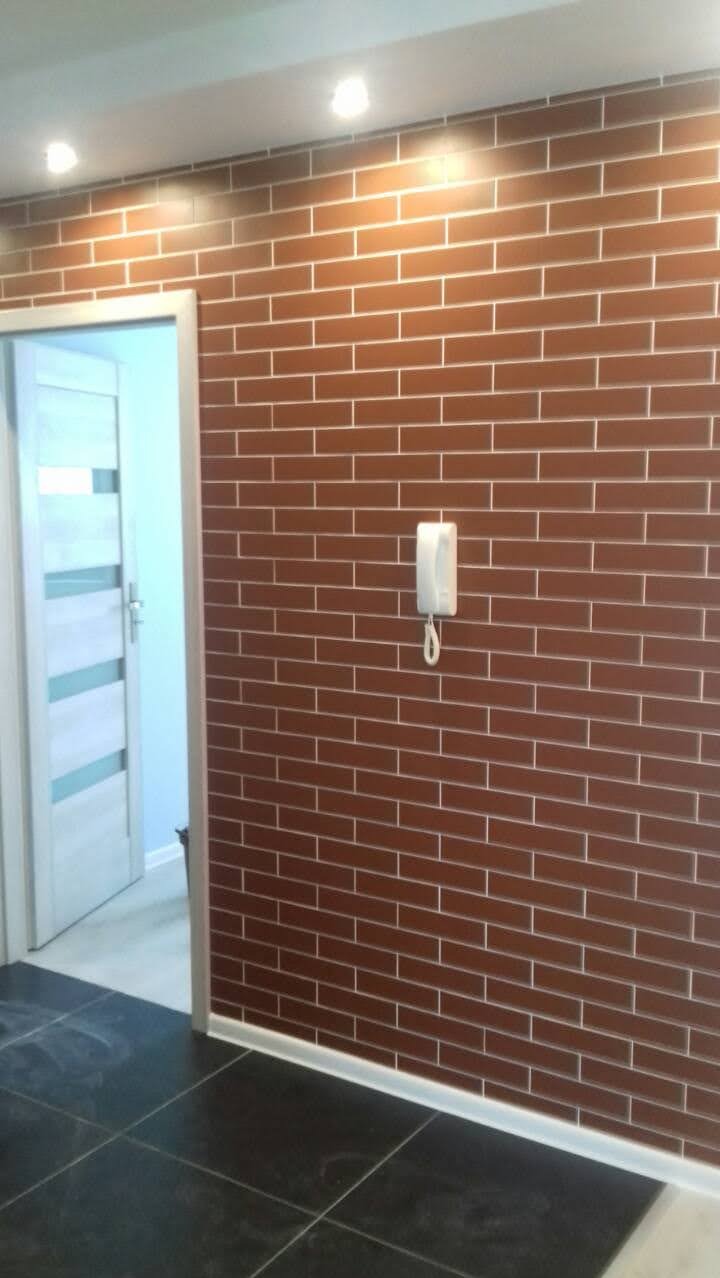 Korytarz z brązową ścianą wyłożoną płytkami imitującymi cegłę, z białymi fugami i zamontowanym domofonem, czarna podłoga z płytek, białe drzwi wewnętrzne z przeszkleniami w tle.