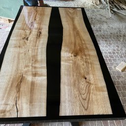 F.H.U. HUBERTUS Hubert Wójtowicz - Blat stołu z dwoma deskami z jasnego drewna oddzielonymi czarną żywicą epoksydową, widoczne sęki i pęknięcia w drewnie, czarna rama stołu, w tle warsztat stolarski z narzędziami i stertą drewna.