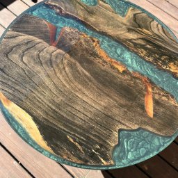 F.H.U. HUBERTUS Hubert Wójtowicz - Okrągły blat stołu z żywicy epoksydowej i drewna o wyraźnych słojach, imitujący rzekę, ułożony na drewnianym tarasie.