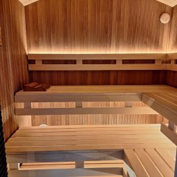 F.H.U. HUBERTUS Hubert Wójtowicz - Wnętrze sauny z drewnianymi ławkami na różnych poziomach, oświetleniem LED podkreślającym pionowy wzór boazerii i zagłówkiem na górnej ławce.