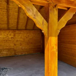 Drewniana altana z beżową podłogą, widok na solidny słup nośny i ściany z desek, oświetlone słońcem. Detal konstrukcyjny, świeże drewno.