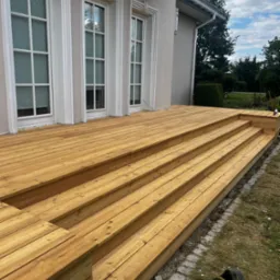 Nowy, drewniany taras z kilkoma stopniami, przylegający do budynku z białymi oknami. Na trawie leży but. Widok na ogród w tle.