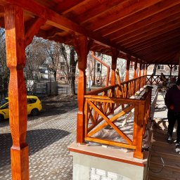 F.H.U. HUBERTUS Hubert Wójtowicz - Zadaszony taras z drewnianą balustradą w kolorze miodowym, widok z zewnątrz. Częściowo wykończony, z widocznymi narzędziami i elementami konstrukcyjnymi. Żółty samochód w tle.