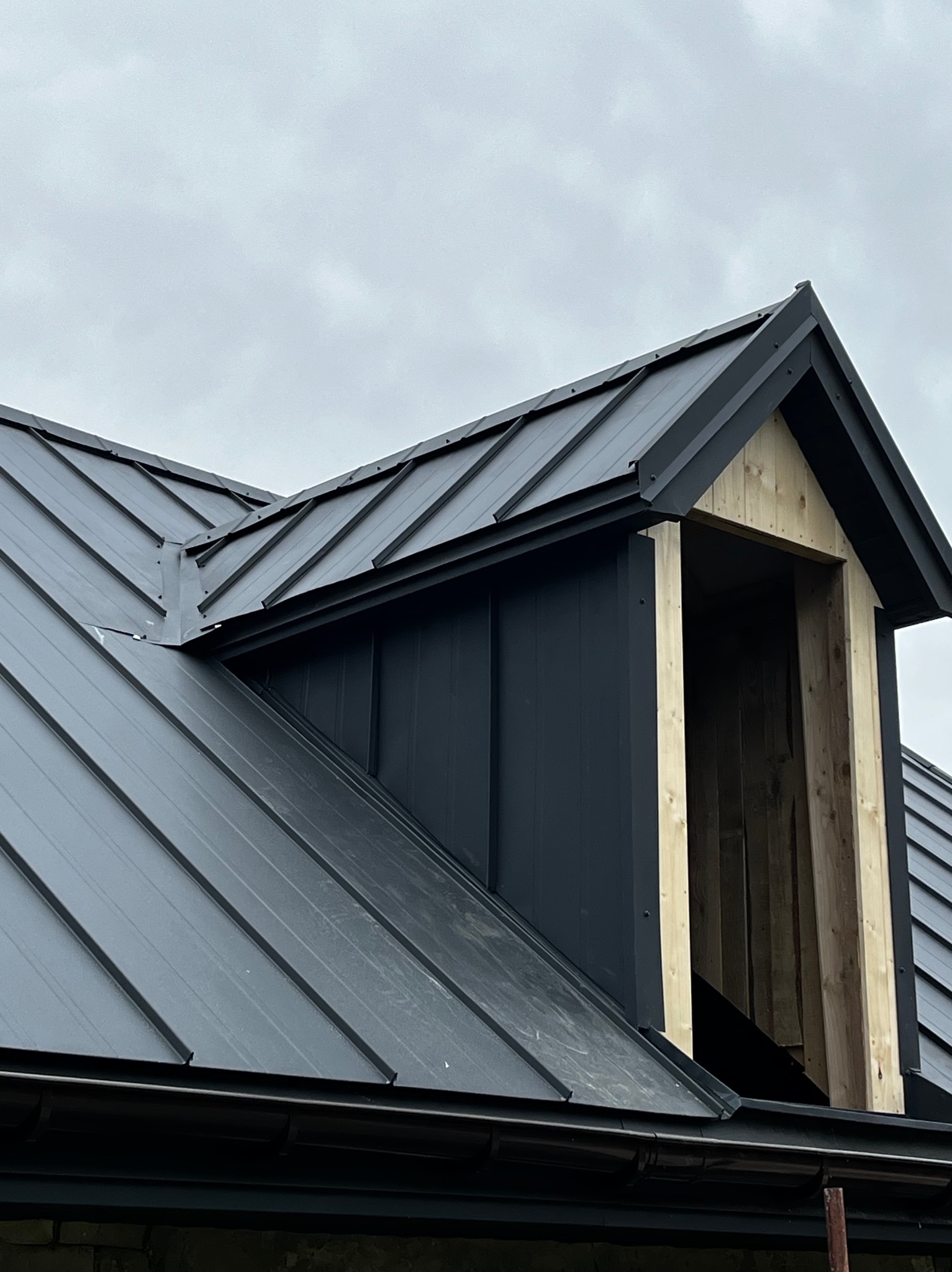 Fragment dachu z blachy na rąbek stojący w kolorze grafitowym, z oknem dachowym w trakcie montażu, widok z dołu na tle pochmurnego nieba.