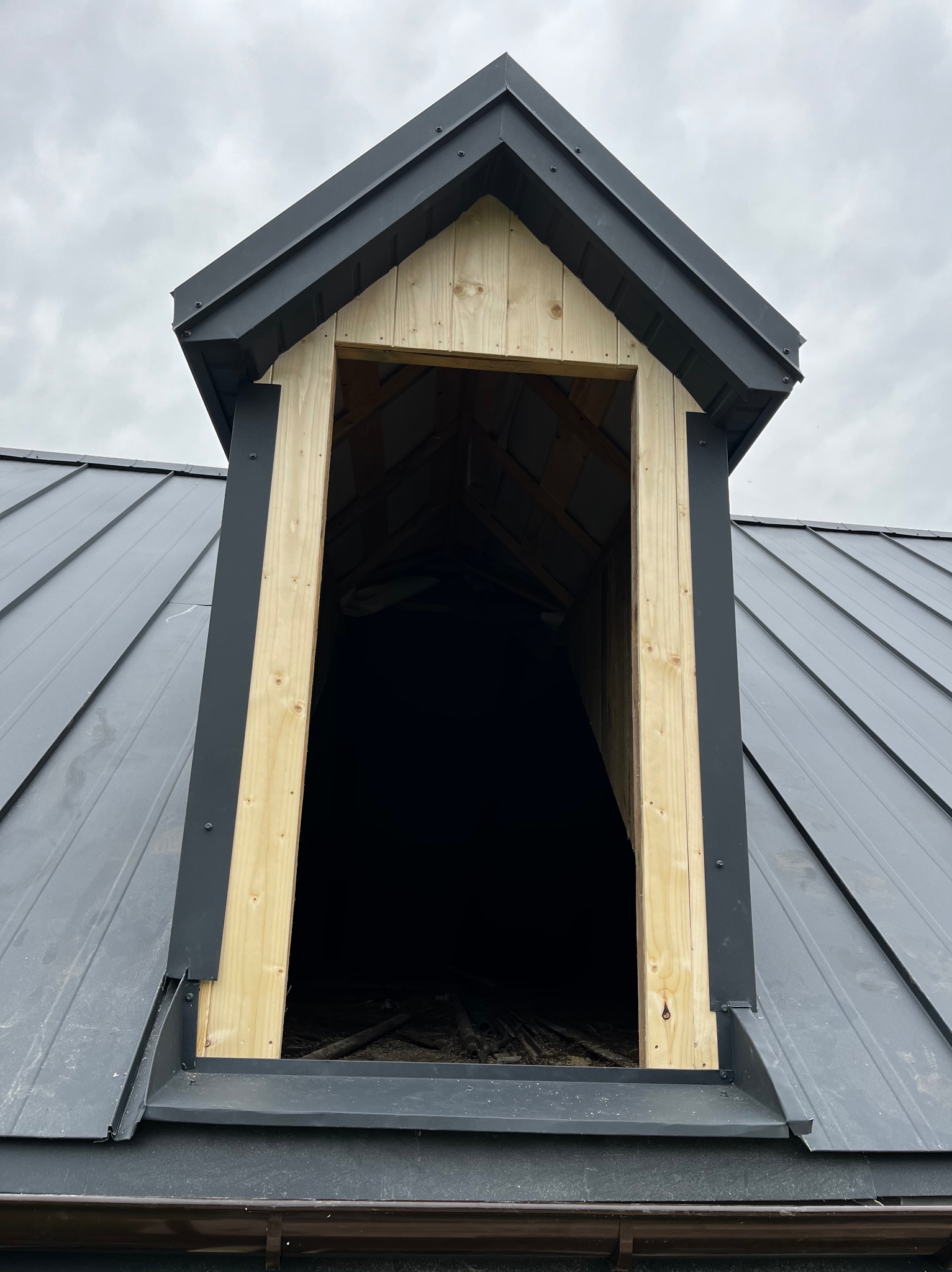 Nowe okno dachowe z drewnianą ramą i czarnymi obróbkami blacharskimi na metalowym dachu w kolorze grafitowym, widok z bliska na tle pochmurnego nieba.