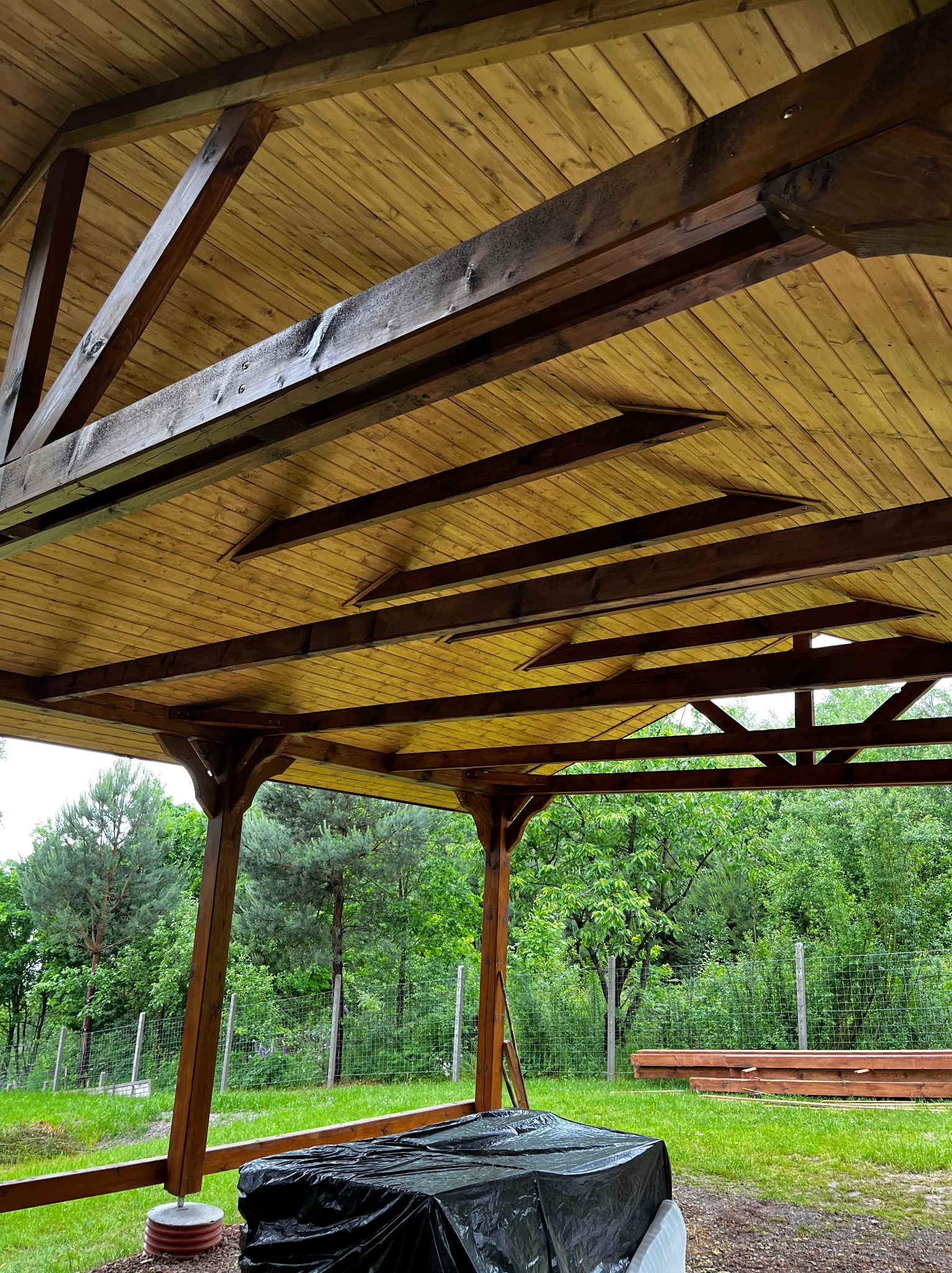 Drewniana altana z widoczną konstrukcją dachu i belkami, w tle zieleń drzew. Część materiałów budowlanych przykryta czarną folią. Widok z wnętrza altany.