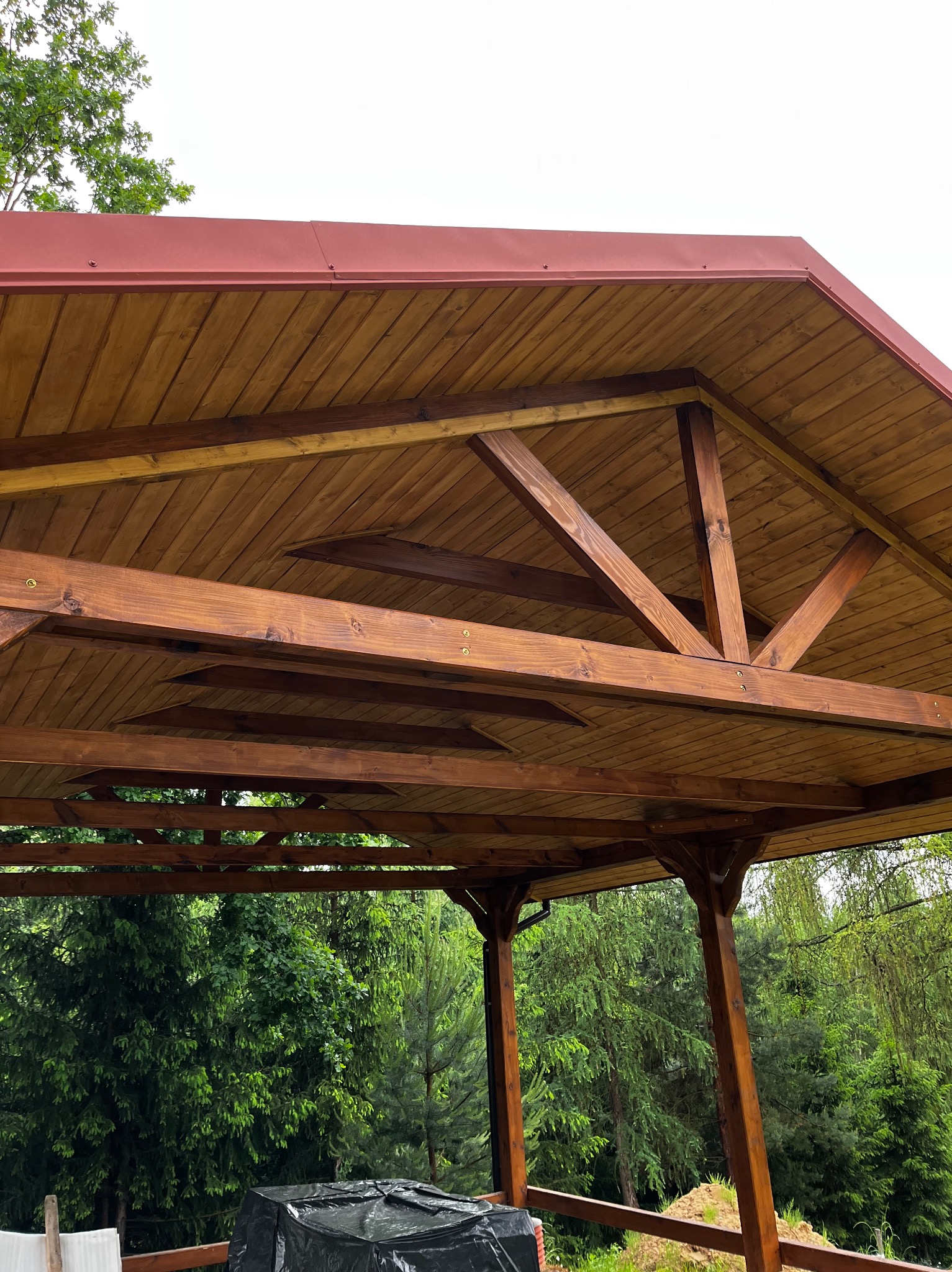 Drewniana altana z czerwonym, metalowym dachem, widok od wewnątrz, z zewnątrz widoczne drzewa. Konstrukcja pomalowana na brązowo, z widoczną strukturą drewna.