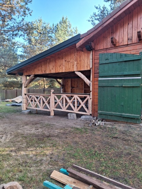 Drewniana altana w trakcie budowy, surowe drewno, widoczne elementy konstrukcyjne, zielone drzwi budynku w tle, ziemiste podłoże. Jasne oświetlenie.