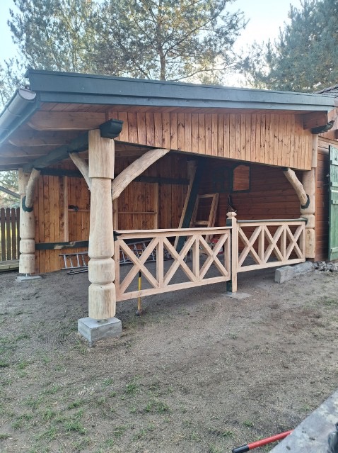 Drewniana altana z ozdobną balustradą i kolumnami na betonowych podstawach. Widoczne elementy konstrukcyjne i zadaszenie. Otoczenie: ziemia, trawa, drzewa.