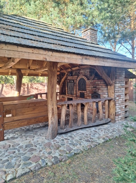 Drewniana altana z ceglanym piecem i kominem, kryta gontem bitumicznym, otoczona kamienną ścieżką i zielenią. Rustykalny styl, naturalne materiały.