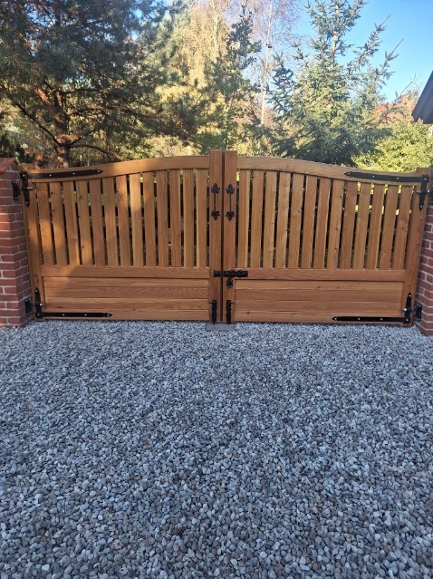 Drewniana brama wjazdowa z dekoracyjnymi zawiasami i zamkiem, osadzona na ceglanych słupkach, na tle zieleni i żwirowego podłoża w słoneczny dzień.