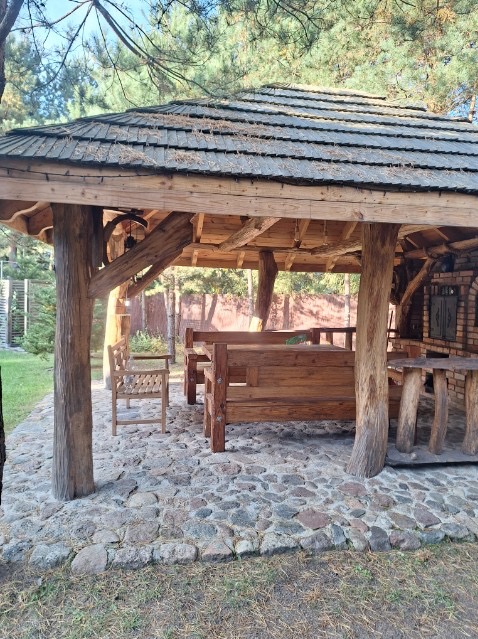 Drewniana altana z dachem krytym gontem, otoczona kamienną nawierzchnią, z drewnianymi meblami wewnątrz i widocznym grillem murowanym z cegły.