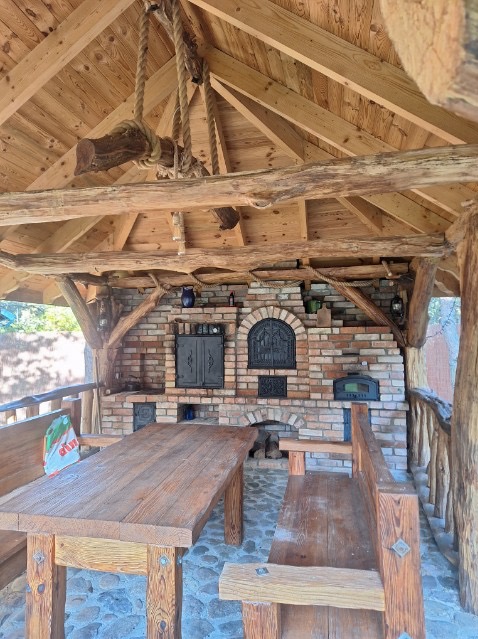 Drewniana altana z murowanym grillem i piecem chlebowym. Solidny stół i ławy z surowego drewna. Dekoracyjne belki stropowe i liny z drewnianym ciężarkiem.