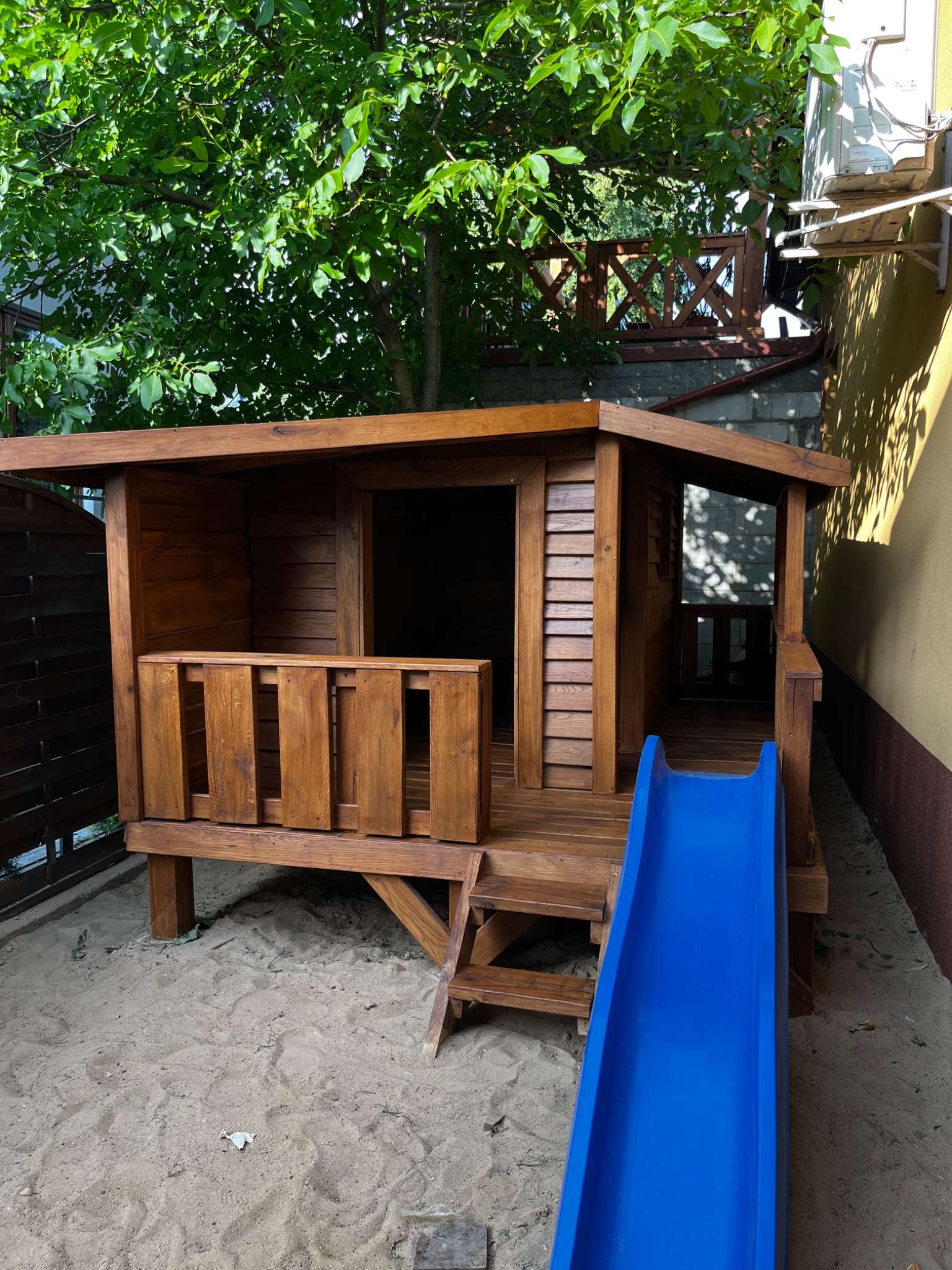 Drewniany domek dla dzieci z niebieską zjeżdżalnią, zbudowany na piasku w ogrodzie. Widoczne schody, zadaszenie i elementy balustrady. Słoneczny dzień.