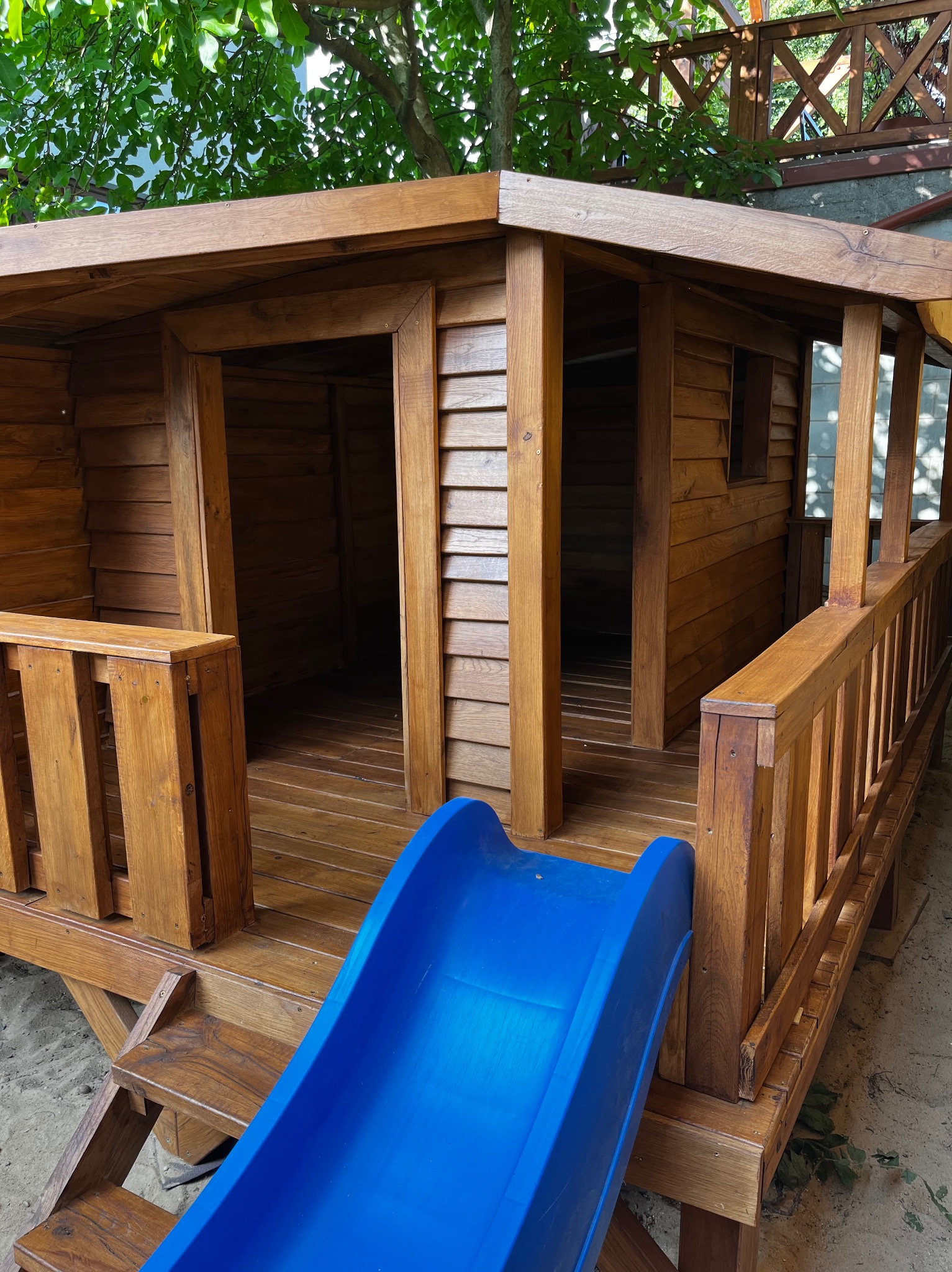 Drewniany domek dla dzieci z niebieską zjeżdżalnią, widok z bliska, otoczony zielenią. Konstrukcja z balustradą i schodkami, elementy drewniane starannie wykończone.