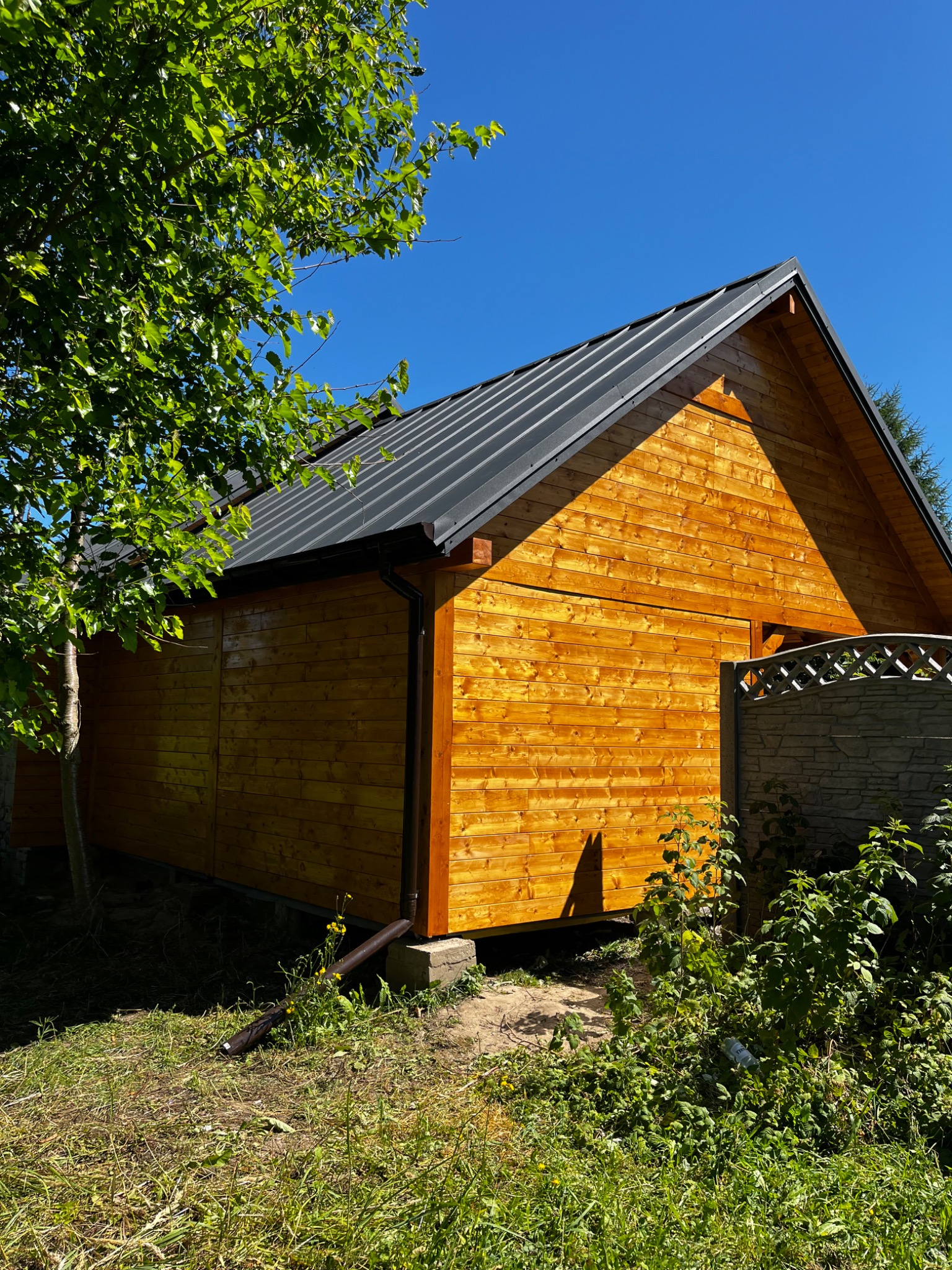 Drewniana altana z ciemnym dachem blachodachówkowym, otoczona zielenią. Elementy obróbki blacharskiej widoczne. Ujęcie z dołu, podkreślające konstrukcję.