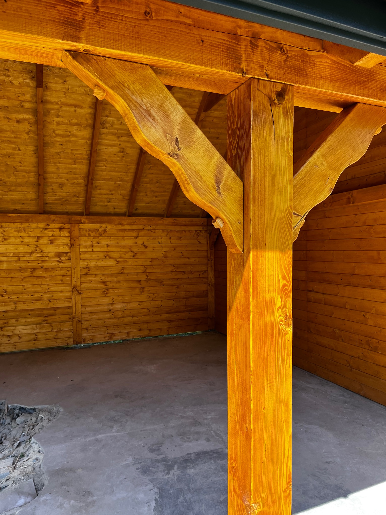 Drewniana altana z beżową podłogą, widok na solidny słup nośny i ściany z desek, oświetlone słońcem. Detal konstrukcyjny, świeże drewno.