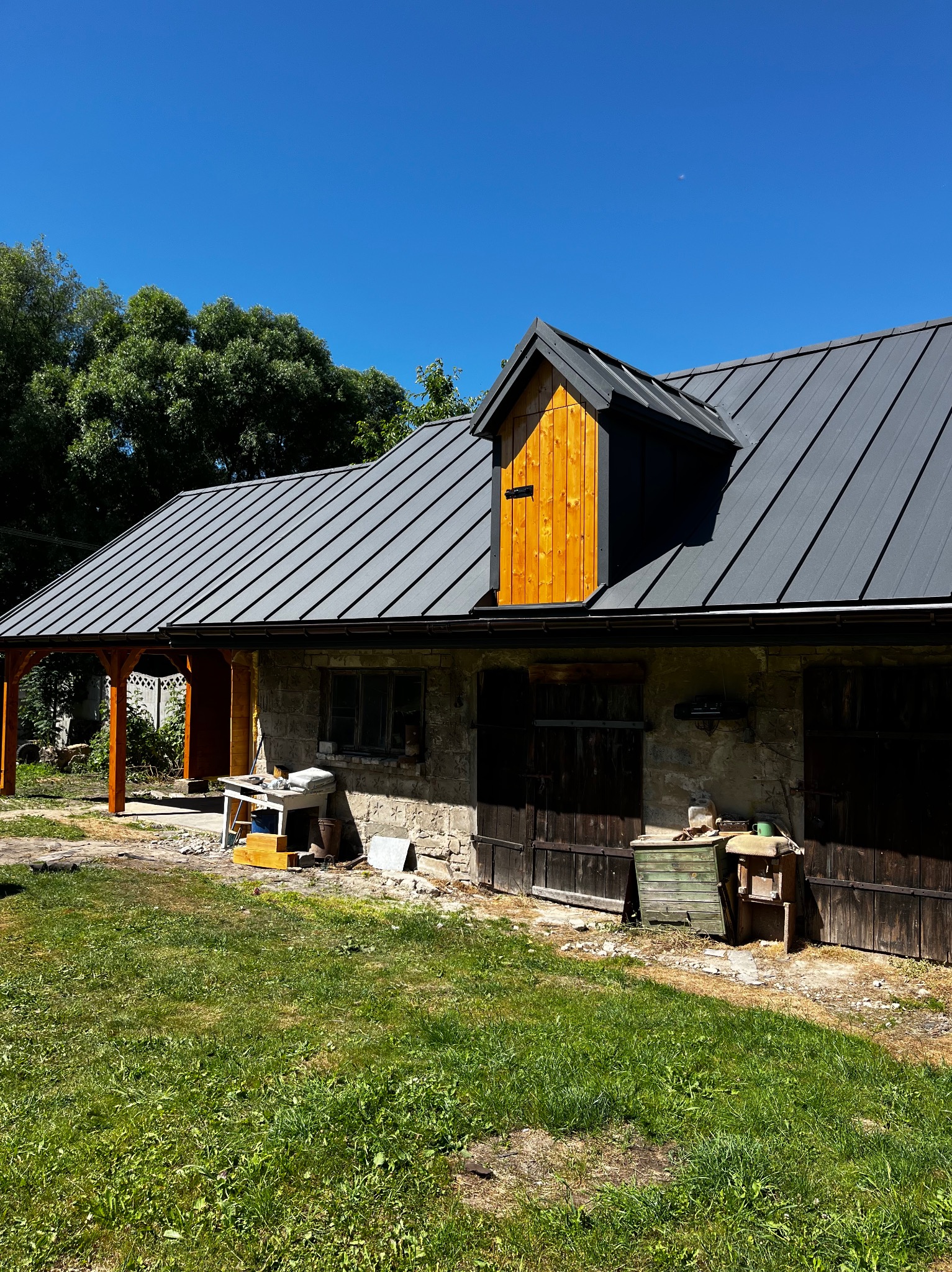 Szary dach z drewnianą lukarną w Bałtowie. Nowe pokrycie dachu na kamiennym budynku. Widoczny fragment drewnianej konstrukcji i narzędzia.