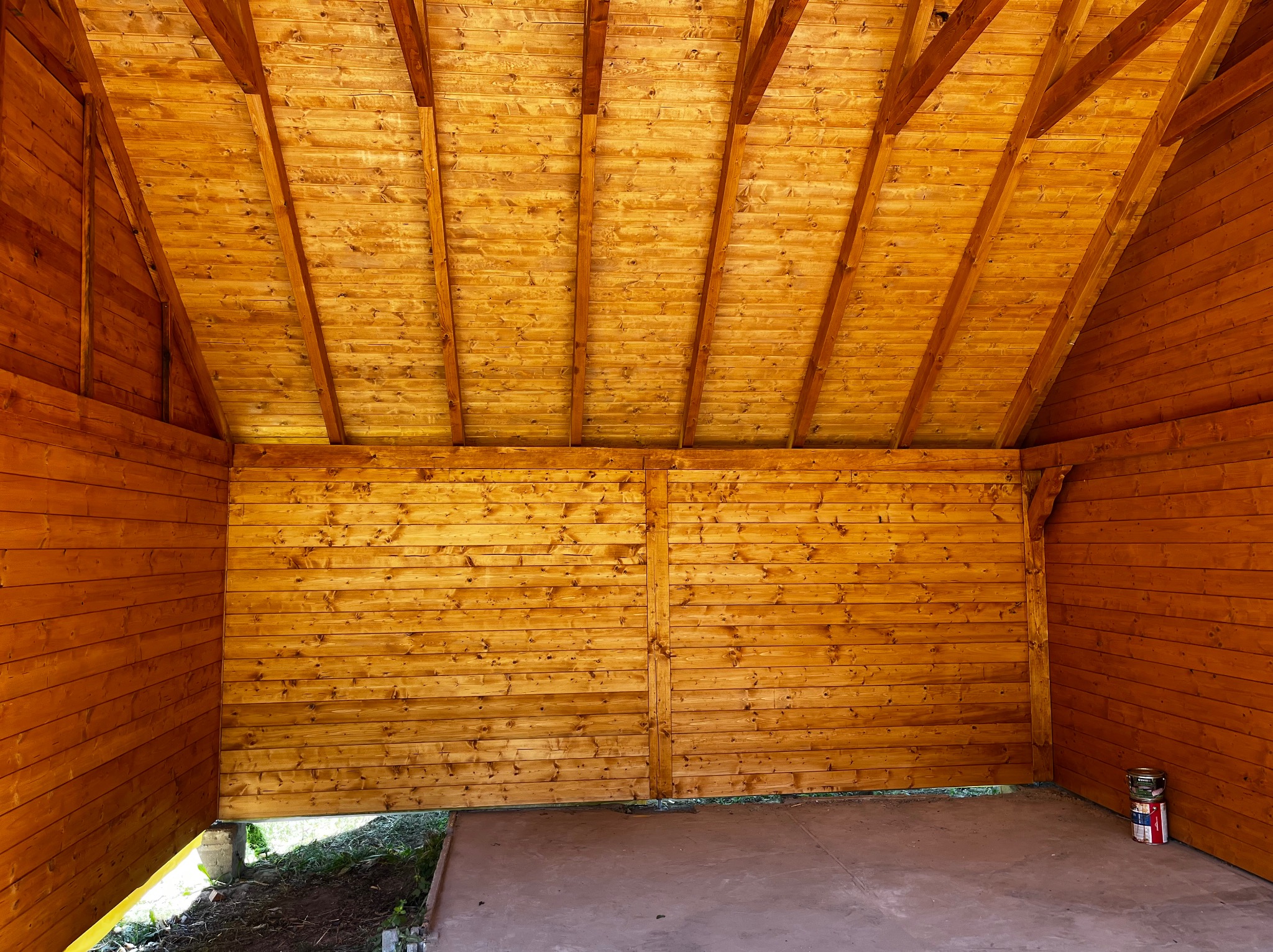 Wnętrze drewnianej altany z widoczną konstrukcją dachu i ścianami z desek, betonowa podłoga, puszki z farbą w rogu. Naturalny kolor drewna.