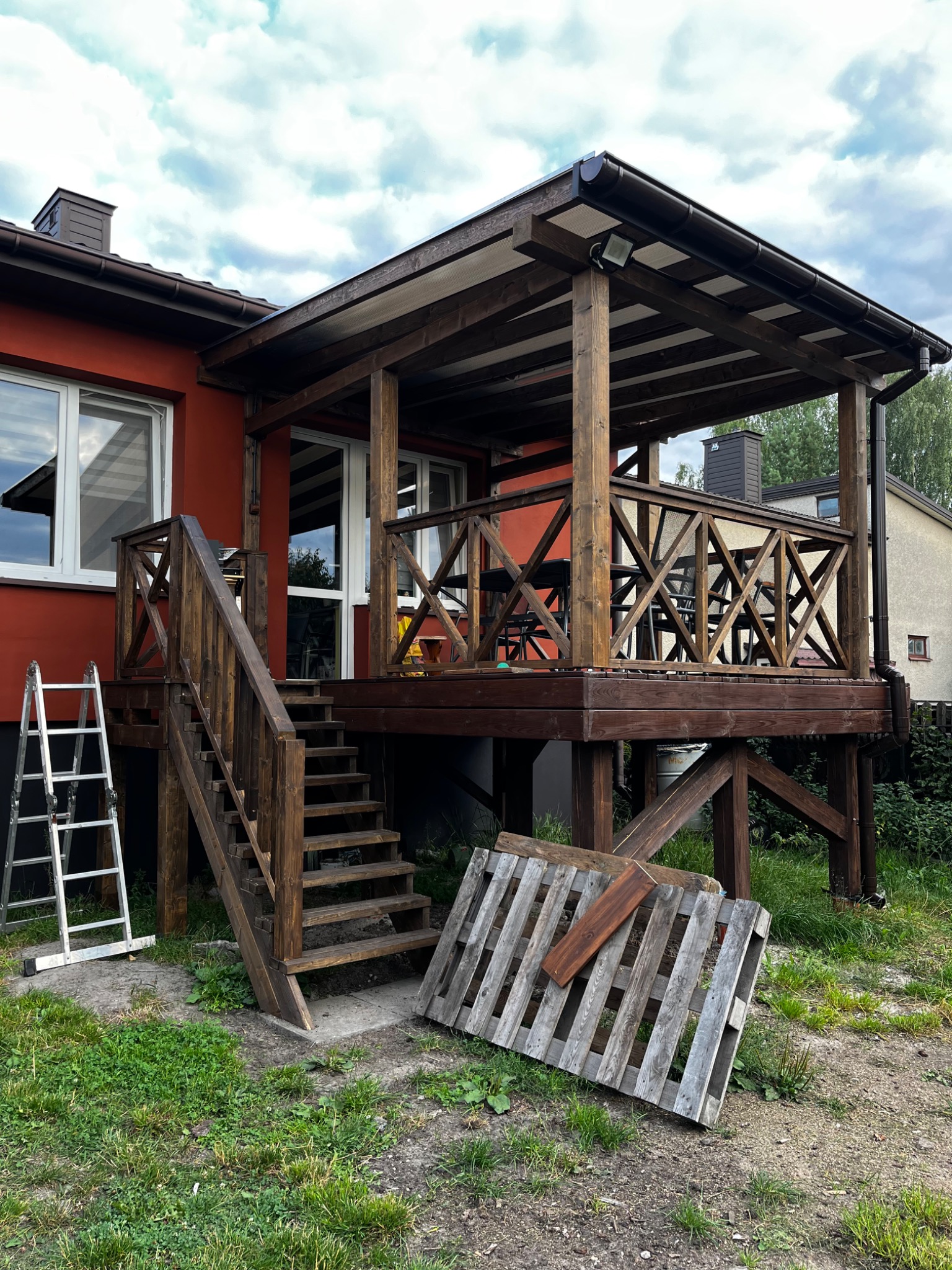 Drewniany taras z zadaszeniem przy domu z czerwonymi ścianami. Schody z balustradą, drabina i paleta obok. Zadaszenie z oświetleniem punktowym.