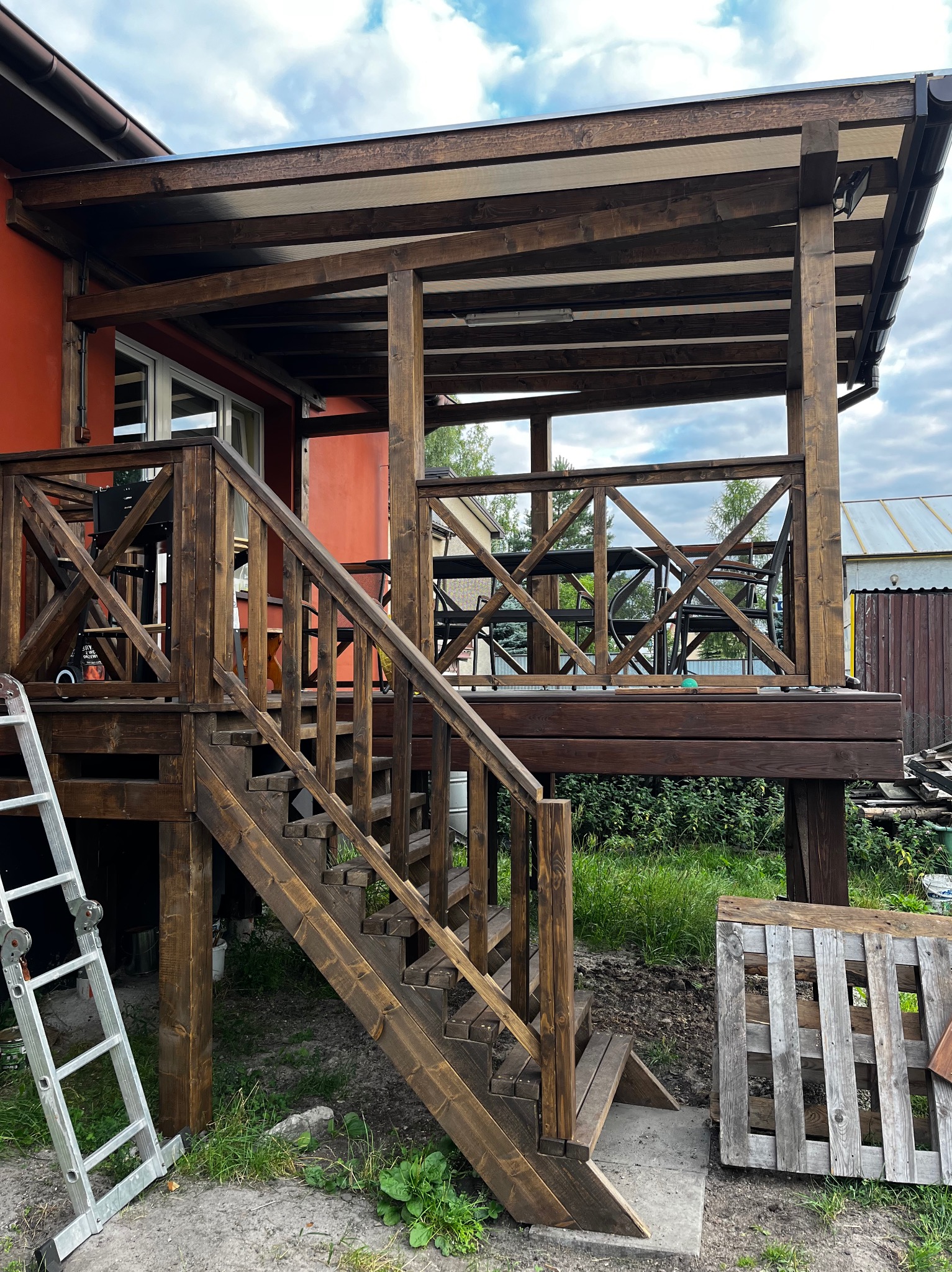 Zadaszony taras drewniany z balustradą krzyżową i schodami, pomalowany na ciemny brąz, z drabiną opartą o bok i paletą obok schodów.
